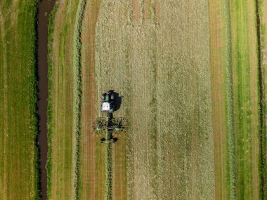 Güneşli bir günde, dar bir su kanalının yanında düzgün sıralarda çim biçen ya da biçen çizgili yeşil ve sarı bir tarlada çalışan bir traktörün hava görüntüsü..