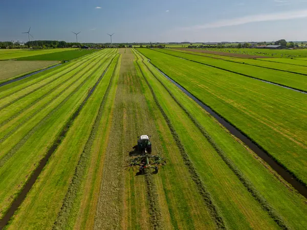 Yeşil arazili Hollanda kırsal manzarası, arazide çalışan bir traktör, çiftlik binaları, rüzgar türbinleri, ve modern tarım ve yenilenebilir enerji sergilenen bir kanal..