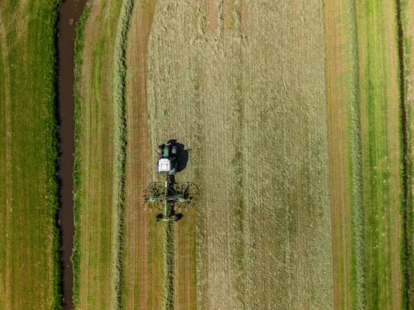 Güneşli bir günde, dar bir su kanalının yanında düzgün sıralarda çim biçen ya da biçen çizgili yeşil ve sarı bir tarlada çalışan bir traktörün hava görüntüsü..