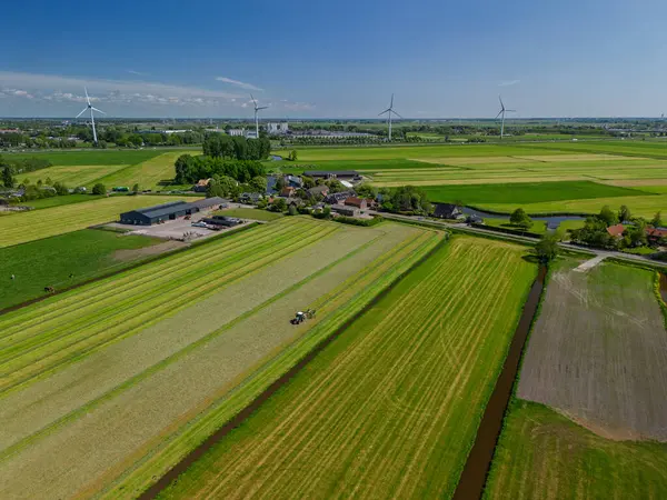 Yeşil arazili Hollanda kırsal manzarası, arazide çalışan bir traktör, çiftlik binaları, rüzgar türbinleri, ve modern tarım ve yenilenebilir enerji sergilenen bir kanal..