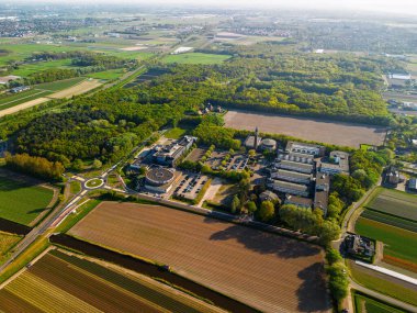 Etrafı tarım arazileri, yeşil ormanlar, bir döner kavşak ve yumuşak güneş ışığı olan açık gökyüzü altındaki uzak kasabalarla çevrili büyük bir bina kompleksi olan kırsal bir bölgenin havadan görünüşü..