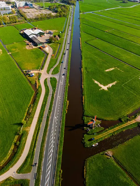 Geleneksel bir yel değirmeni olan Hollanda kırsalının havadan görünüşü. Kanal, yeşil tarlalar ve açık mavi gökyüzü altında hareket eden arabaların olduğu yakın bir yol..
