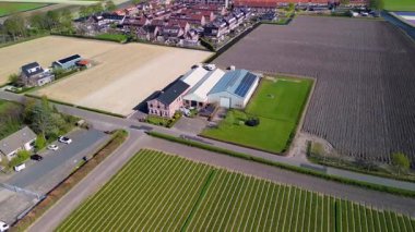 Temiz gökyüzü altında modern evleri ve tarım binalarını harmanlayan tarım arazileri, seralar ve yerleşim alanları olan huzurlu bir kırsal bölge gösteren hava videosu..