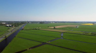 Geleneksel bir yel değirmeni olan Hollanda kırsalının havadan görünüşü. Kanal, yeşil tarlalar ve açık mavi gökyüzü altında hareket eden arabaların olduğu yakın bir yol..