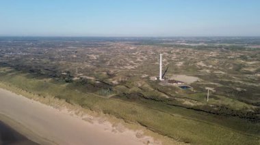 Kum tepeleri ve açık mavi gökyüzü ve sakin deniz ile çevrili sahil boyunca uzanan ve sakin doğal atmosfer yaratan çarpıcı bir kumlu sahil manzarası. Uzun iletişim kulesi