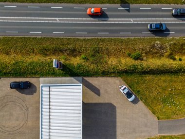 Hareket halindeki arabaların olduğu bir otobanın tepeden aşağı havadan görüntüsü, park etmiş araçların olduğu bir park alanı, beyaz çatılı bir bina, ve yolu ve park yerini ayıran çimenli bir arazi şeridi..