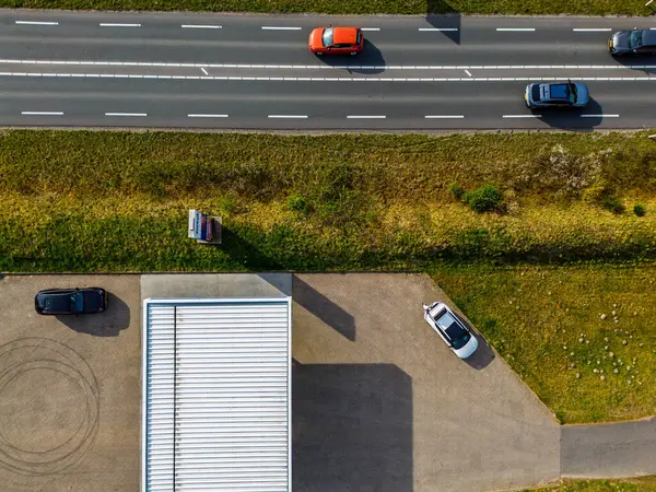 Hareket halindeki arabaların olduğu bir otobanın tepeden aşağı havadan görüntüsü, park etmiş araçların olduğu bir park alanı, beyaz çatılı bir bina, ve yolu ve park yerini ayıran çimenli bir arazi şeridi..