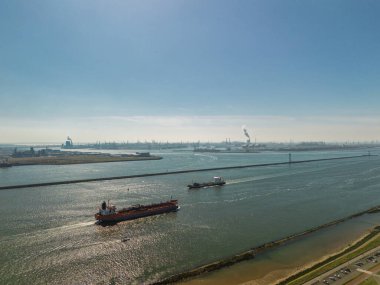 Endüstriyel bir liman yakınındaki geniş bir nehir veya kanalda ilerleyen kargo gemilerinin hava görüntüsü açık mavi gökyüzünün altında vinçler ve bacalar var..