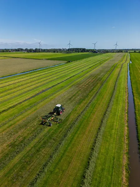 Yeşil arazili Hollanda kırsal manzarası, arazide çalışan bir traktör, çiftlik binaları, rüzgar türbinleri, ve modern tarım ve yenilenebilir enerji sergilenen bir kanal..