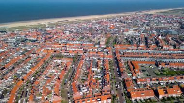 Kırmızı çatılı evleri, spor sahaları, yeşil alanları ve açık mavi gökyüzünün altındaki sahili gösteren bir sahil manzarası..