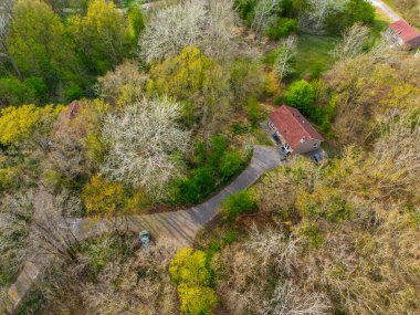 Kırmızı kiremitli çatısı olan küçük bir evin tepeden aşağı havadan görünüşü bahar başlarında ağaçlarla çevrili bir orman ve park halindeki bir arabaya giden dar bir garaj yolu..