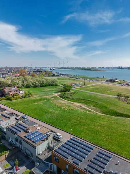 Yeşil parkları, yerleşim alanları, nehir yolu ve işlek nakliye yolları olan bir sahil kasabasının havadan görüntüsü. Rüzgar türbinleri ve endüstriyel liman tesisleri su üzerinde görülebilir..