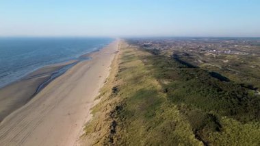 Yumuşak dalgaların kumlu sahille buluştuğu sahilin havadan görünüşü. İnsanlar kuma dağılmış durumda, bazıları kumdan kaleler yapıyor, diğerleri güneşli günün tadını çıkarıyor..
