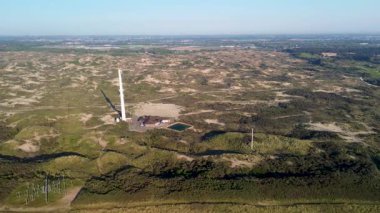 Kum tepeleri ve açık mavi gökyüzü ve sakin deniz ile çevrili sahil boyunca uzanan ve sakin doğal atmosfer yaratan çarpıcı bir kumlu sahil manzarası. Uzun iletişim kulesi