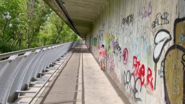 Bratislava, Bratislava Bölgesi, Slovakya Cumhuriyeti - 09.07.2024: Bir çocuk, yeşil ağaçlarla çevrili modern bir yaya köprüsünde, güpegündüz grafiti kaplı bir duvar boyunca yürüyor.