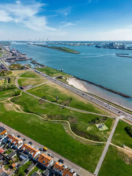 Yeşil parkları, yerleşim alanları, nehir yolu ve işlek nakliye yolları olan bir sahil kasabasının havadan görüntüsü. Rüzgar türbinleri ve endüstriyel liman tesisleri su üzerinde görülebilir..