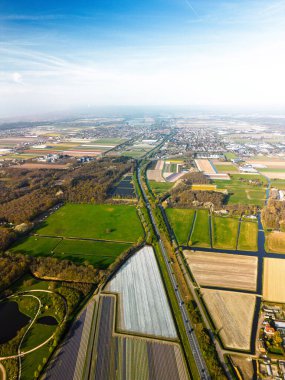 Hollanda kırsalının hava panoramik manzarasında tarım arazileri, yeşil tarlalar, ormanlar ve uzak bir kasabaya doğru uzanan bir otoyol var..