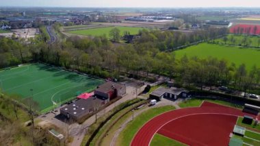 Yeşil futbol sahası ve kırmızı atletizm pisti olan, ağaçlar ve kırsal bölgelerle çevrili, çiçek tarlaları ve tarlaları olan bir spor kompleksinin havadan görünüşü..