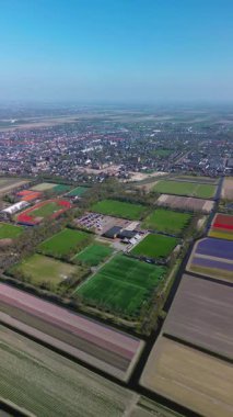 Birçok futbol sahası, bir pist ve saha stadyumu, tenis kortu ve park alanı olan, yemyeşil orman ve renkli lale tarlalarıyla çevrili bir spor kompleksinin havadan görüntüsü..