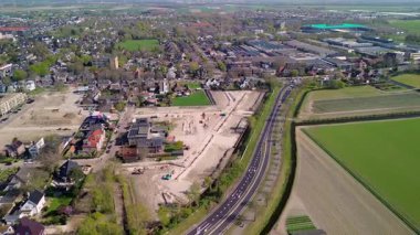 Hava görüntüleri, inşaat alanları, konut binaları, ana yol ve çevresindeki yeşil tarım alanlarının bulunduğu bir banliyö bölgesini gösteriyor..