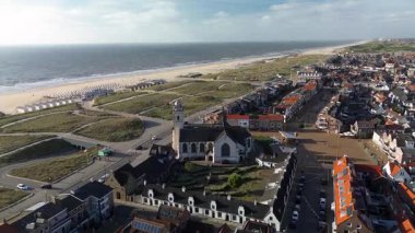 Beyaz bir kilise, kırmızı çatılı evler, dolambaçlı yollar ve Kuzey Denizi kıyısı boyunca sahil kulübeleri olan geniş kumlu bir sahil manzarası..