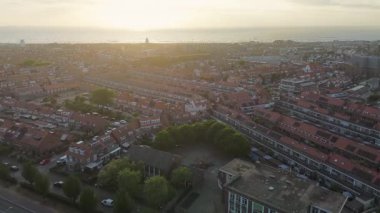 Hollanda kıyı kasabasının üzerinde gün batımı manzarası. Kırmızı çatılı evler, ağaçlarla kaplı sokaklar ve ufukta görünen deniz, altın ışığın altında..