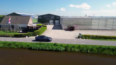 Renkli bir lale tarlasının çiçeklerin arasında yürüyen insansız hava aracı görüntüsü. Siyah bir araba, güneşli bir bahar gününde bir kanal ve modern bir evin yanında kırsal bir yol boyunca sürer..