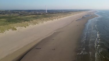 Kum tepeleri ve açık mavi gökyüzü ve sakin deniz ile çevrili sahil boyunca uzanan ve sakin doğal atmosfer yaratan çarpıcı bir kumlu sahil manzarası. Uzun iletişim kulesi