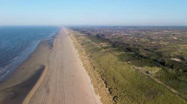 Sahil kulübeleriyle dolu güzel bir kumsal manzarası, kıyıda dinlenen insanlar ve ön planda çimenli kum tepeleriyle çevrili, kıyıya vuran nazik dalgalar..