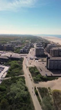 Daire binaları, kum tepeleri ve açık mavi gökyüzünün altında geniş kumlu bir plaj olan modern bir sahil manzarası. Yolda yürüyen insanlar, bisikletçiler ve arabalar var..