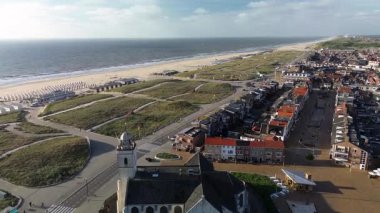 Hollanda kıyı kasabasının üzerinde gün batımı manzarası. Kırmızı çatılı evler, ağaçlarla kaplı sokaklar ve ufukta görünen deniz, altın ışığın altında..