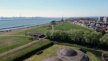 Sahil manzaralı sakin bir sahil kasabası kayalık bir kıyı şeridi, yeşil açık alanlar, geçen araçların olduğu bir yol ve açık mavi gökyüzünün altındaki konut binalarını gösteriyor. Rüzgar Değirmenleri.