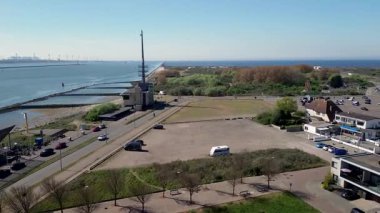 Sahil manzaralı sakin bir sahil kasabası kayalık bir kıyı şeridi, yeşil açık alanlar, geçen araçların olduğu bir yol ve açık mavi gökyüzünün altındaki konut binalarını gösteriyor. Rüzgar Değirmenleri.