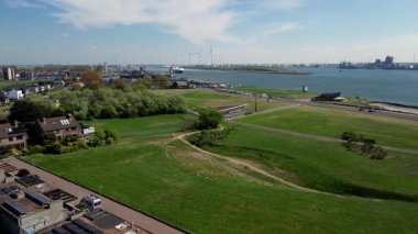 Yeşil parkları, yerleşim alanları, nehir yolu ve işlek nakliye yolları olan bir sahil kasabasının havadan görüntüsü. Rüzgar türbinleri ve endüstriyel liman tesisleri su üzerinde görülebilir..