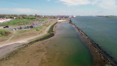 Denize doğru uzanan dalgalı bir dalgakıranın olduğu kıyı şeridi uzaktaki rüzgar türbinleriyle kaplı. Bir sahil, gezinti güvertesi ve yeşil hendek boyunca uzanan küçük binalar..