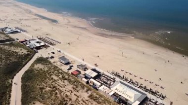 Sahil kulübeleriyle dolu güzel bir kumsal manzarası, kıyıda dinlenen insanlar ve ön planda çimenli kum tepeleriyle çevrili, kıyıya vuran nazik dalgalar..