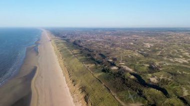 Sahil kulübeleriyle dolu güzel bir kumsal manzarası, kıyıda dinlenen insanlar ve ön planda çimenli kum tepeleriyle çevrili, kıyıya vuran nazik dalgalar..