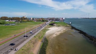 Denize doğru uzanan dalgalı bir dalgakıranın olduğu kıyı şeridi uzaktaki rüzgar türbinleriyle kaplı. Bir sahil, gezinti güvertesi ve yeşil hendek boyunca uzanan küçük binalar..