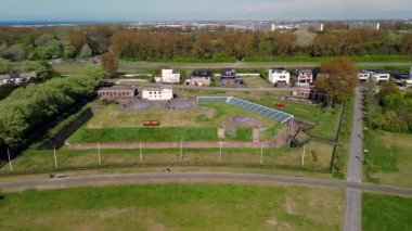 Modern binalar, ağaçlar ve yürüyüş yolları ile çevrili çimenlerle kaplı tarihi bir askeri kalenin havadan görünüşü, kıyı şeridi yakınlarında parlak mavi bir gökyüzünün altında..