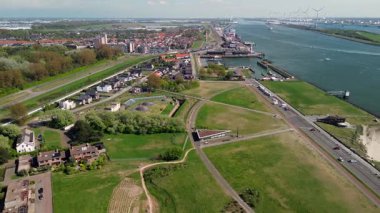 Yeşil parkları, yerleşim alanları, nehir yolu ve işlek nakliye yolları olan bir sahil kasabasının havadan görüntüsü. Rüzgar türbinleri ve endüstriyel liman tesisleri su üzerinde görülebilir..