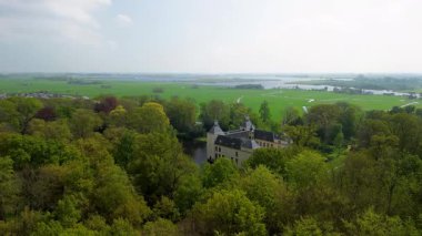 Uçsuz bucaksız yeşil tarlaları, dolambaçlı su yolları ve Hollanda 'nın düz kırsalında parçalı bulutlu bir gökyüzünün altında uzanan bir nehirle çevrili tarihi bir şatonun havadan görünüşü..