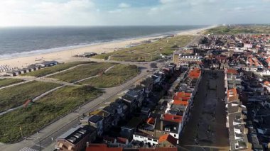 Hollanda kıyı kasabasının üzerinde gün batımı manzarası. Kırmızı çatılı evler, ağaçlarla kaplı sokaklar ve ufukta görünen deniz, altın ışığın altında..