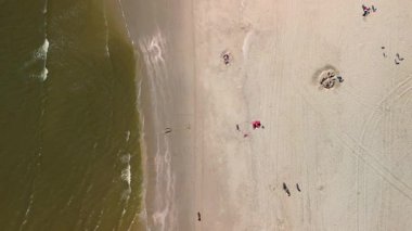 Sahil kulübeleriyle dolu güzel bir kumsal manzarası, kıyıda dinlenen insanlar ve ön planda çimenli kum tepeleriyle çevrili, kıyıya vuran nazik dalgalar..