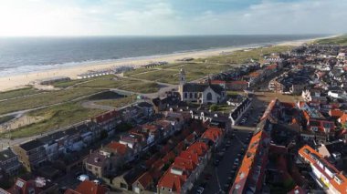 Hollanda kıyı kasabasının üzerinde gün batımı manzarası. Kırmızı çatılı evler, ağaçlarla kaplı sokaklar ve ufukta görünen deniz, altın ışığın altında..