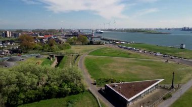 Yeşil parkları, yerleşim alanları, nehir yolu ve işlek nakliye yolları olan bir sahil kasabasının havadan görüntüsü. Rüzgar türbinleri ve endüstriyel liman tesisleri su üzerinde görülebilir..