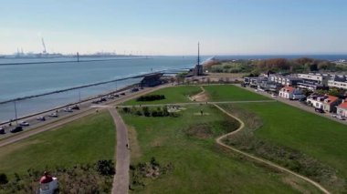 Sahil manzaralı sakin bir sahil kasabası kayalık bir kıyı şeridi, yeşil açık alanlar, geçen araçların olduğu bir yol ve açık mavi gökyüzünün altındaki konut binaları..