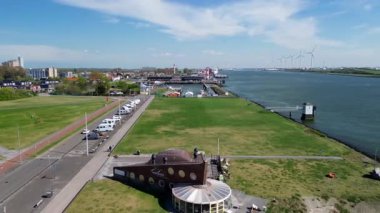 Modern bir binayla güneşli bir nehir kenarı manzarası, sıra sıra karavanlar ve uzakta bir liman. Rüzgar türbinleri açık mavi gökyüzünün altında ufukta döner.