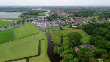 Dar bir su kanalı çevreleyen ağaçları yansıtan yeşil bir ormandan geçiyor. Arka planda güzel bir Hollanda köyü var. Yatı olan büyük bir iskele. Yeşil bir çayır, bir tarla.