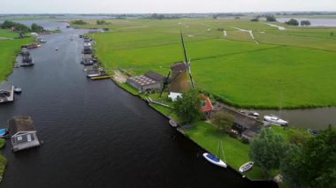 Bereketli yeşil tarlalar boyunca uzanan dar bir kanalın havadan görünüşü ve berrak mavi gökyüzünün altında limanı ve demirlemiş yatları olan huzurlu bir Hollanda köyüne uzanan küçük bir orman. Güzel bir rüzgar değirmeni.