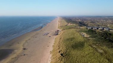 Sahil kulübeleriyle dolu güzel bir kumsal manzarası, kıyıda dinlenen insanlar ve ön planda çimenli kum tepeleriyle çevrili, kıyıya vuran nazik dalgalar..